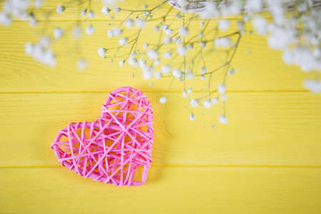 Pink wicker heart and fresh bouquet of white gypsophila on yellow wooden background. Spring romantic card with empty space.の写真素材