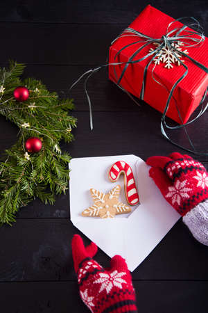 Hands in red winter gloves holding white envelope with Christmas gingerbread on black wooden background. New Year homemade cookie with fir wreath and gift box.の写真素材