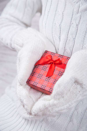 Hands in white knitted mittens holding red gift box. Christmas present. New Year card.の写真素材