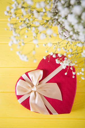 Gift box in shape of heart and fresh bouquet of white gypsophila on yellow wooden background. Spring romantic present for women with flowers.の写真素材