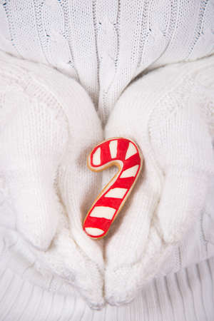 Hands in white knitted mittens holding Christmas gingerbread in form of caramel cane. New Year homemade cookie.の写真素材