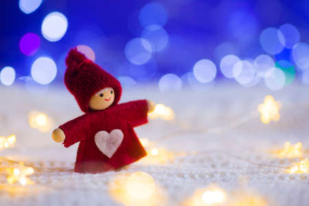 Christmas little red doll with heart surrounded with warm garland lights and blue bokeh on knitted white fabric. St. Valentine day romantic card with copy space.の写真素材