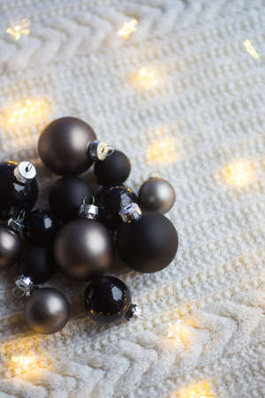 Metallic and black Christmas balls surrounded with warm garland lights on knitted white fabric in daylight. Stylish New Year decor at home.の写真素材