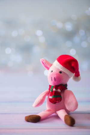 Closeup cute toy pig in Santa hat and scarf sitting on wooden table on light bokeh background. Symbol of Chinese New Year.の写真素材