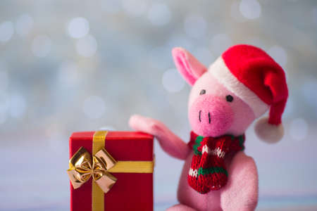 Closeup cute toy pig in Santa hat and scarf sitting on wooden table and holding festive gift box on light bokeh background. Symbol of Chinese New Year.の写真素材