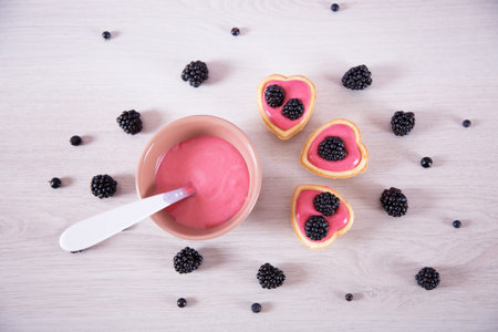 Pink berry yogurt decorated with blackberries in bowl and heart-shaped tartlets, top view. Tender...
