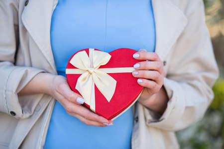 Young female hands holding gift box in shape of red heart with bow. Romantic gift for woman.の写真素材