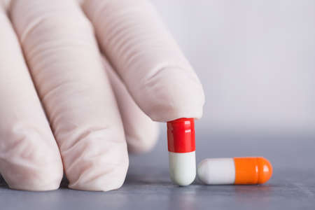 Closeup hand in medical latex glove holding two different colorful capsules on gray table.の写真素材