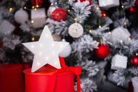 White glowing LED star on red gift box near decorated Christmas tree. New Year festive atmosphere.の写真素材