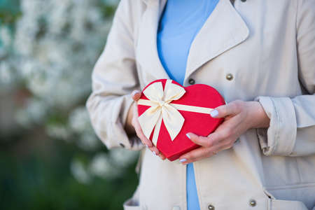 Young female hands holding gift box in shape of red heart with blurred spring background. Romantic gift for woman.の写真素材