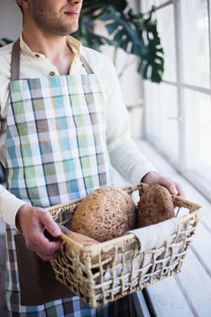 Brutal baker holding basket with freshly baked bread of different kinds with bran in his hands near window. Home bakery.の写真素材