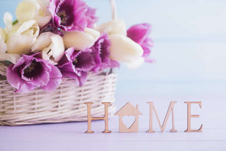Wooden inscription "Home" and bouquet of tender tulips in wicker basket on pastel background. Spring composition.の写真素材
