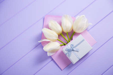 Beautiful tender bouquet of white tulips in envelope with gift box on lilac wooden background. Spring composition. Card for women or mother day.の写真素材