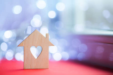 Wooden icon of house with hole in the form of heart on red table near window in daylight with bokeh lights on blurred background. Romantic card with copy space. Concept of sweet home.の写真素材