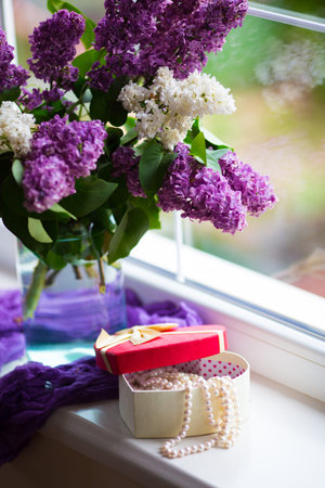 Heart shaped gift box with pearl necklace and tender bouquet of beautiful lilac in glass vase near window in daylight. Spring blooming flowers with romantic present for woman.の写真素材