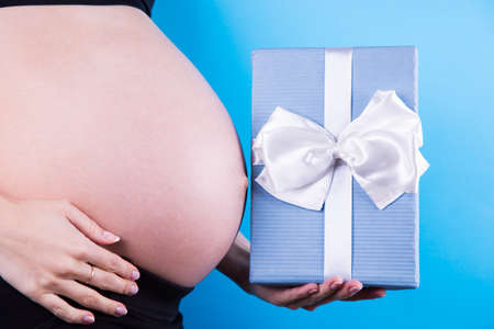 Pregnant woman is holding festive gift box in her hand near belly on blue background. Concept of baby surprise, suddenly pregnancy. Present for mother. Studio shot with copy space.の写真素材