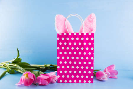 Toy Easter bunny ears sticking out of spotted paper shopping bag with pink tulips on blue background. Concept of spring Easter gift.の写真素材