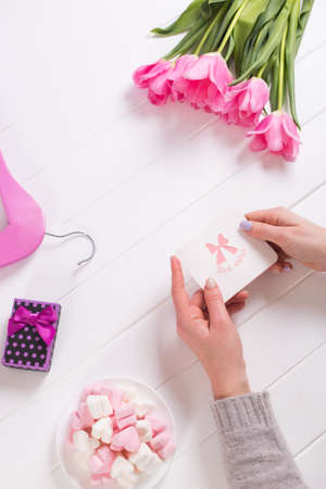 Young female hands holding greeting card near bouquet of pink tulips, gift box, hanger and marshmallow. Spring gift for woman or mother with best wishes.の写真素材