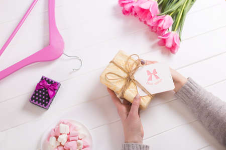 Young female hands holding gift with greeting card near bouquet of pink tulips, hanger and marshmallow. Spring gift for woman or mother with best wishes.の写真素材