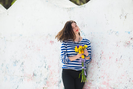 Summer young woman in stripped vest with vibrant bouquet of yellow freesias and irises near light...