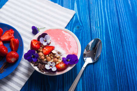 Pink yogurt bowl with strawberry, nuts, chocolate, decorated with flowers, with spoon on navy wooden background, top view. Spring healthy breakfast, overhead view with copy space.の写真素材