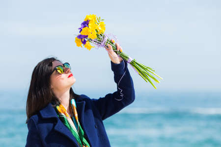 Stylish young woman in sunglasses and coat holding bouquet of freesias and irises with sea landscape on blurred background. Vivid colorful image on sunny day with copy spaceの写真素材
