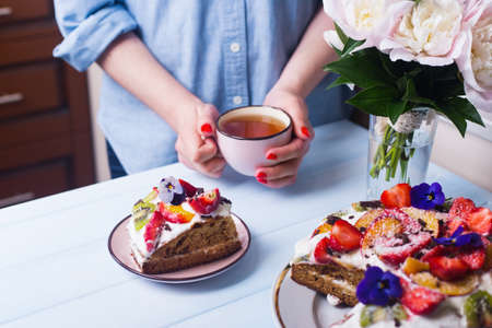 Young female hands holding cup of tea near plate with sour cream cake with fruits decorated with flowers. Spring homemade dessert with kiwi, peach and strawberry.の写真素材