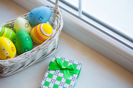 Green gift box and set of decorated colorful Easter eggs in wicker basket near window in daylight. Spring home vibrant composition with copy space.の写真素材