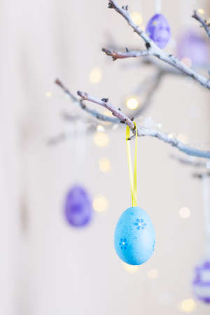 Decorated colorful Easter egg hanging on tree branch indoor with warm bokeh on white background. Spring home decor with garland lights, copy space.の写真素材