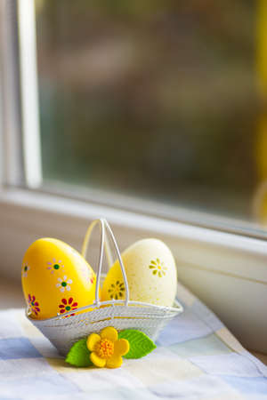Colorful orange and yellow Easter eggs in basket with decorative flower near window in daylight. Spring home vibrant composition with copy space.の写真素材