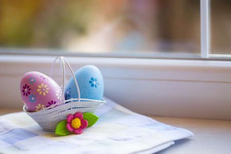 Colorful pink and blue Easter eggs in basket with decorative flower near window in daylight. Spring home vibrant composition with copy space.の写真素材