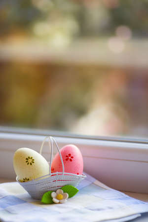 Colorful yellow and pink Easter eggs in basket with decorative flower near window in daylight. Spring home vibrant composition with copy space.の写真素材