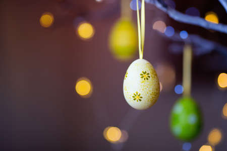 Decorated colorful Easter egg hanging on tree branch indoor with warm bokeh on background. Spring home decor with garland lights, copy space.の写真素材
