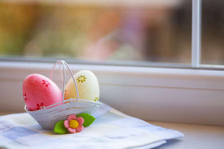 Colorful pink and yellow Easter eggs in basket with decorative flower near window in daylight. Spring home vibrant composition with copy space.の写真素材