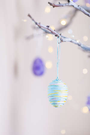 Decorated colorful Easter egg hanging on tree branch indoor with warm bokeh on white background. Spring home decor with garland lights, copy space.の写真素材