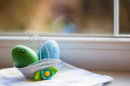 Colorful blue and green Easter eggs in basket with decorative flower near window in daylight. Spring home vibrant composition with copy space.の写真素材