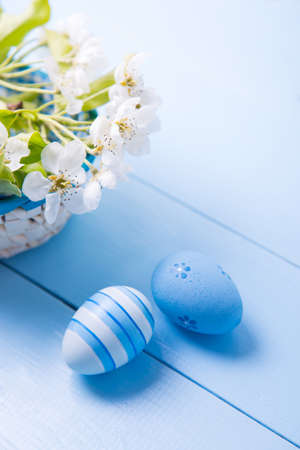 Two painted blue Easter eggs near basket with white spring flowering branch on light blue background.の写真素材