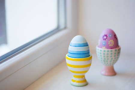 Two colorful painted Easter eggs in vibrant modern egg stands near window in daylight. Spring home minimalistic composition with copy space.の写真素材