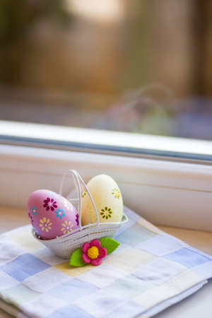 Colorful pink and yellow Easter eggs in basket with decorative flower near window in daylight. Spring home vibrant composition with copy space.の写真素材