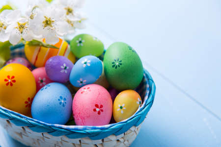 Variety of decorated colorful Easter eggs in basket with white spring flowers on light blue background.の写真素材