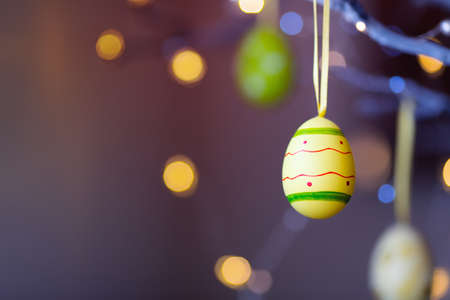 Decorated colorful Easter egg hanging on tree branch indoor with warm bokeh on background. Spring home decor with garland lights, copy space.の写真素材