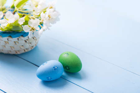 Two painted blue and green Easter eggs near basket with white spring flowering branch on light blue background.の写真素材