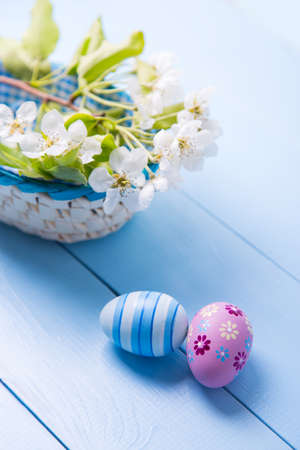 Two painted blue and purple Easter eggs near basket with white spring flowering branch on light blue background.の写真素材