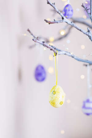 Decorated colorful Easter egg hanging on tree branch indoor with warm bokeh on white background. Spring home decor with garland lights, copy space.の写真素材