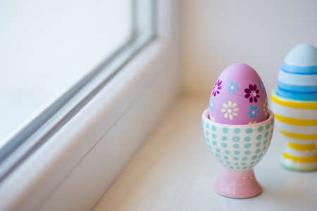Two colorful painted Easter eggs in vibrant modern egg stands near window in daylight. Spring home minimalistic composition with copy space.の写真素材