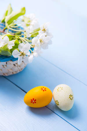Two painted orange and yellow Easter eggs near basket with white spring flowering branch on light blue background.の写真素材