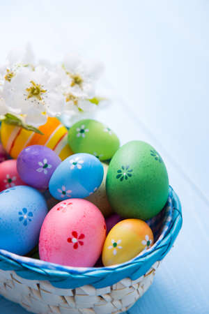 Variety of decorated colorful Easter eggs in basket with white spring flowers on light blue background.の写真素材