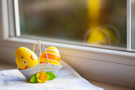 Colorful orange Easter eggs in basket with decorative flower near window in daylight. Spring home vibrant composition with copy space.の写真素材