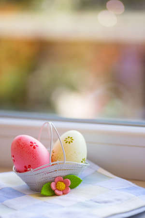 Colorful pink and yellow Easter eggs in basket with decorative flower near window in daylight. Spring home vibrant composition with copy space.の写真素材