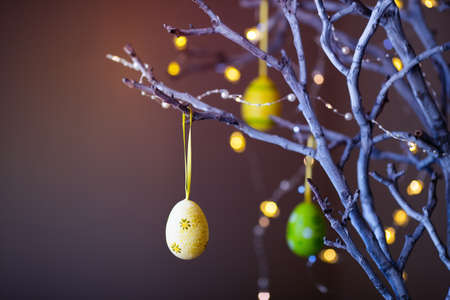 Decorated colorful Easter egg hanging on tree branch indoor with warm bokeh on background. Spring home decor with garland lights, copy space.の写真素材
